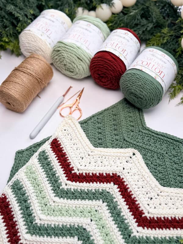 Several skeins of yarn, crochet hooks, and scissors are arranged around a green, red, and white crochet chevron washcloths on a white surface with greenery in the background.