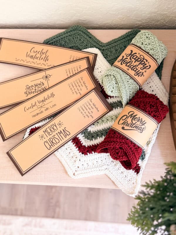 Two crocheted blankets and two rolls of yarn with festive "Merry Christmas" labels are displayed on a wooden surface beside four brown handmade item tags.
