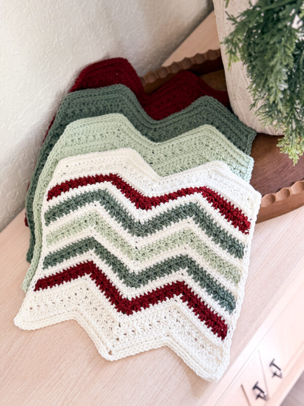 A crocheted chevron-patterned blanket in white, green, and red is folded on a light wood table next to a plant.