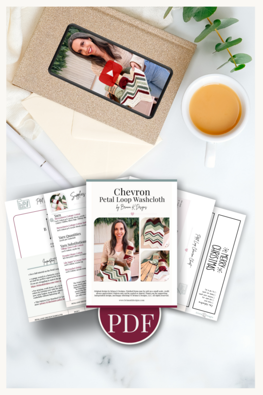 Flat lay of a crochet washcloth PDF pattern, instructional pages, a video thumbnail, a cup of tea, pen, green branch, and white cloth on a light background.