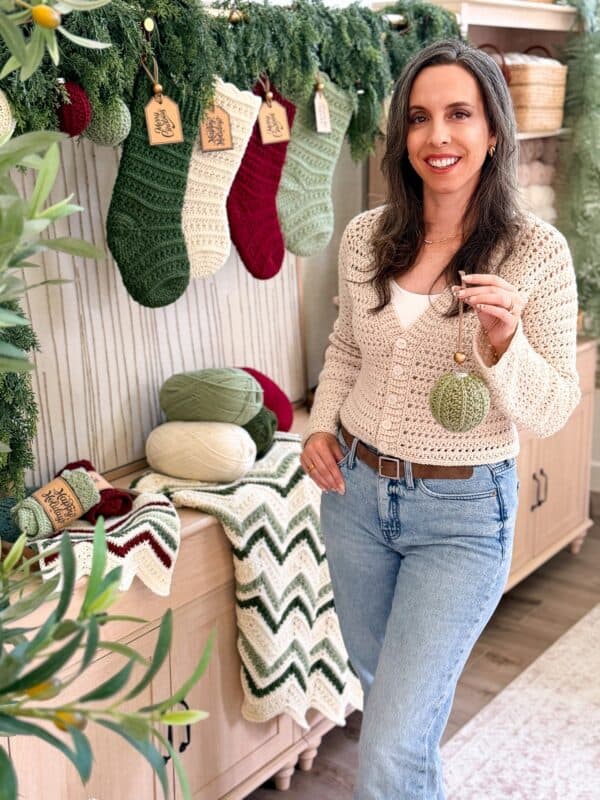 A woman stands indoors holding a crocheted ornament. Behind her are crocheted stockings, yarn, and a chevron-patterned blanket, all arranged as holiday decor.