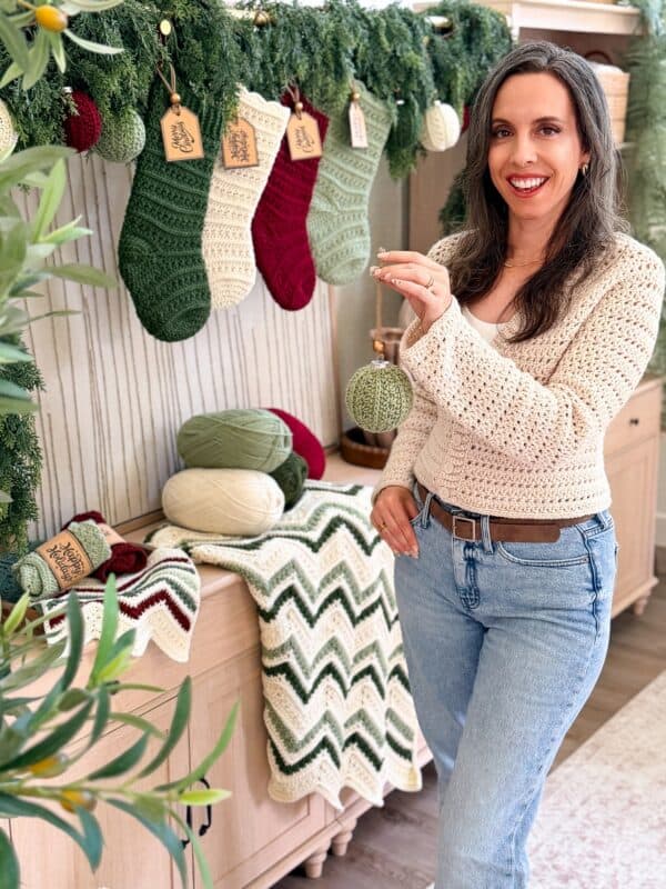 Woman in a cream crochet sweater stands beside a holiday display with stockings, yarn, and a zigzag blanket, holding a green ornament crafted from a crochet ornament pattern.