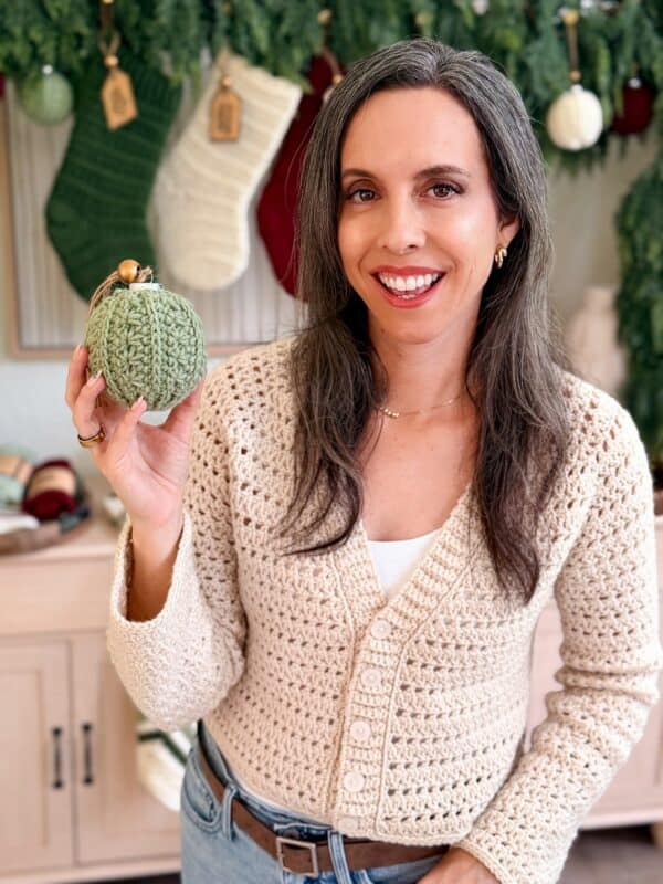 A woman wearing a cream crochet cardigan holds a green crocheted ornament. Holiday stockings and decorations hang in the background.