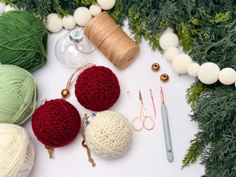 Yarn, twine, crochet hook, scissors, glass ornaments, and crocheted balls are arranged on a white surface with pine branches and a wooden bead garland.