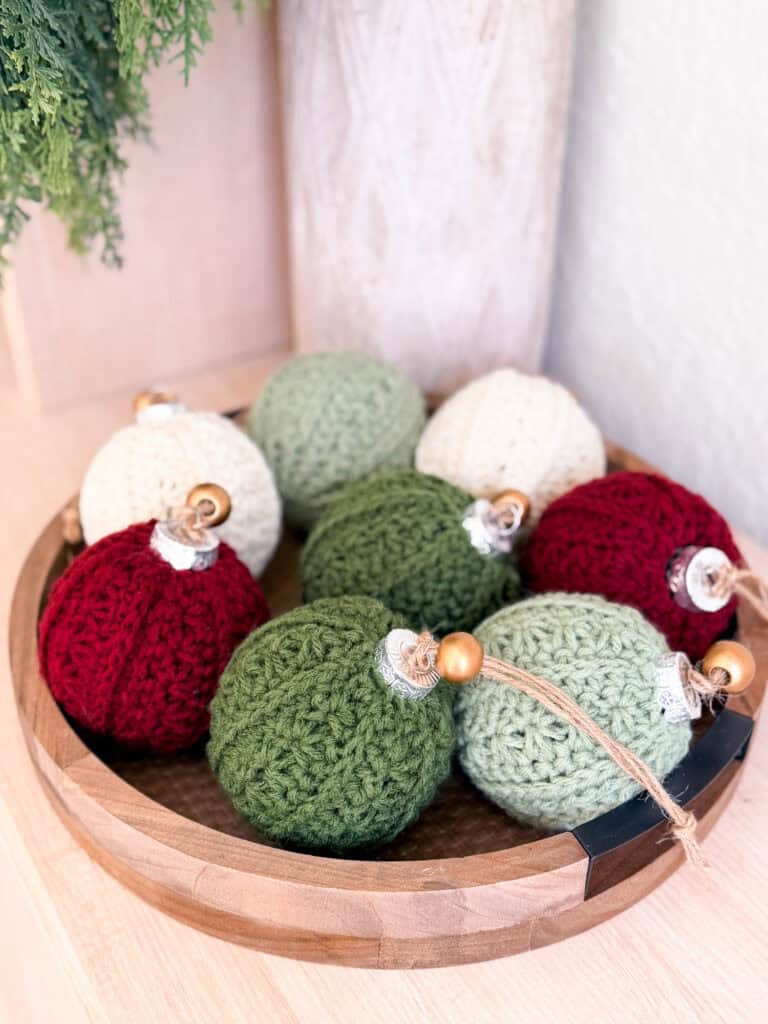 A wooden tray holds eight knitted Christmas ornaments in red, green, and cream colors, each with a wooden bead and string for hanging.