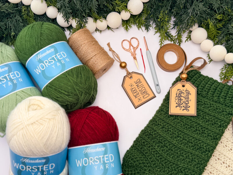 Balls of yarn, crochet tools, twine, tags reading "Merry Christmas" and "Happy Holidays," and a crocheted green item are arranged on a white surface with greenery and garland.