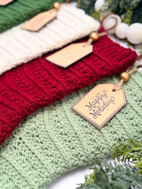 Three knitted holiday stockings in green, red, and white are arranged close together, each with a tag reading "Happy Holidays" and surrounded by greenery.