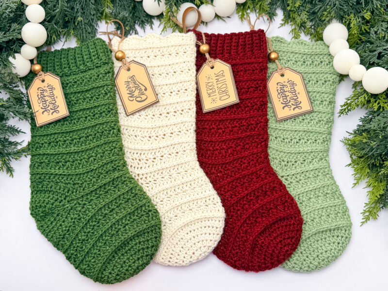 Four crocheted Christmas stockings—green, cream, red, and light green—hang with wooden tags among greenery and white bead garland, each beautifully crafted from a crochet Christmas stocking pattern.