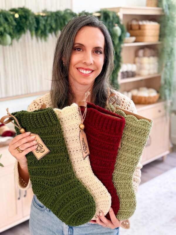 Woman holding three crocheted Christmas stockings in green, cream, and red, standing in a room with yarn and festive decorations.