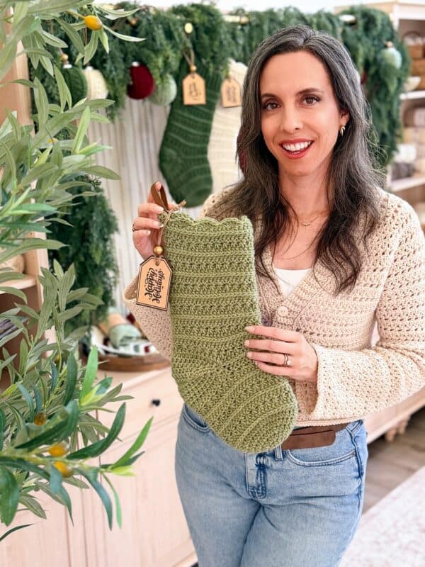 A woman in a cream cardigan holds up a green crocheted stocking with a tag, standing in front of a festive, decorated display with holiday stockings.