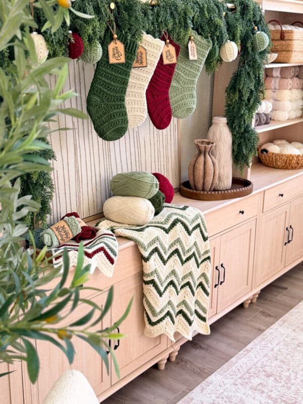 A wooden sideboard decorated with green garland, crochet stockings, yarn, and a chevron-patterned crochet blanket, with vases and neatly stacked towels on top and shelves below.