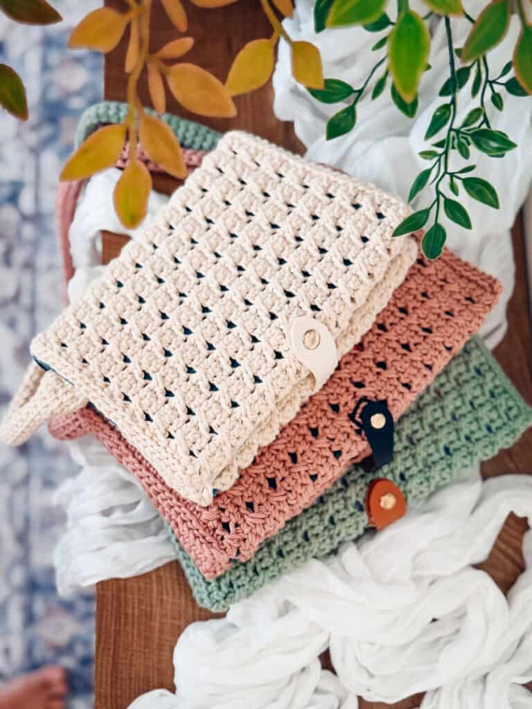 Three crocheted pouches in beige, pink, and green with button closures stacked on a wooden surface, with leafy branches and white fabric nearby.
