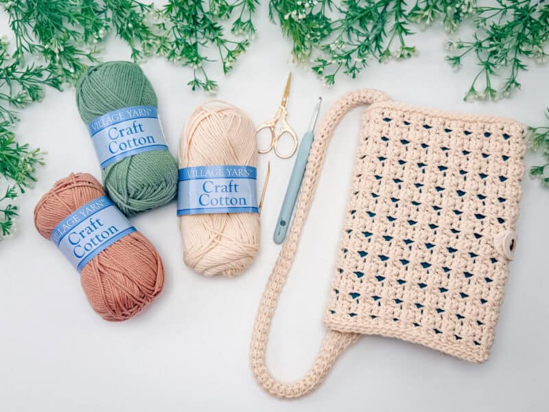 Three skeins of craft cotton yarn, a pair of scissors, a crochet hook, and a crocheted bag are arranged on a white surface with leafy greenery around the edges.