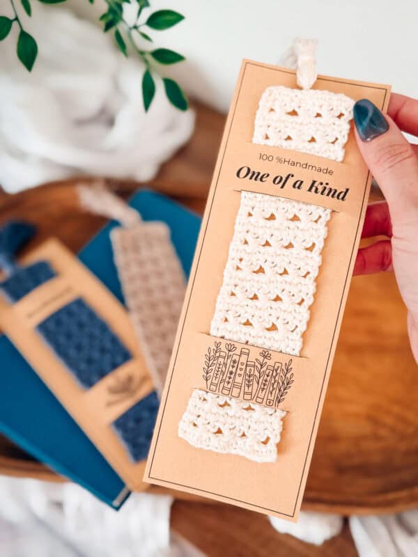 A hand holds a packaged crocheted bookmark labeled "100% Handmade, One of a kind." Two other bookmarks rest on blue books in the background.