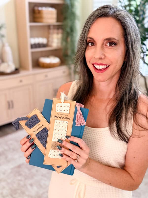 A woman holds three blue books, each with a handmade crochet bookmark attached. The bookmarks have labels reading "Bookworm" and "One of a Kind." She is smiling at the camera.