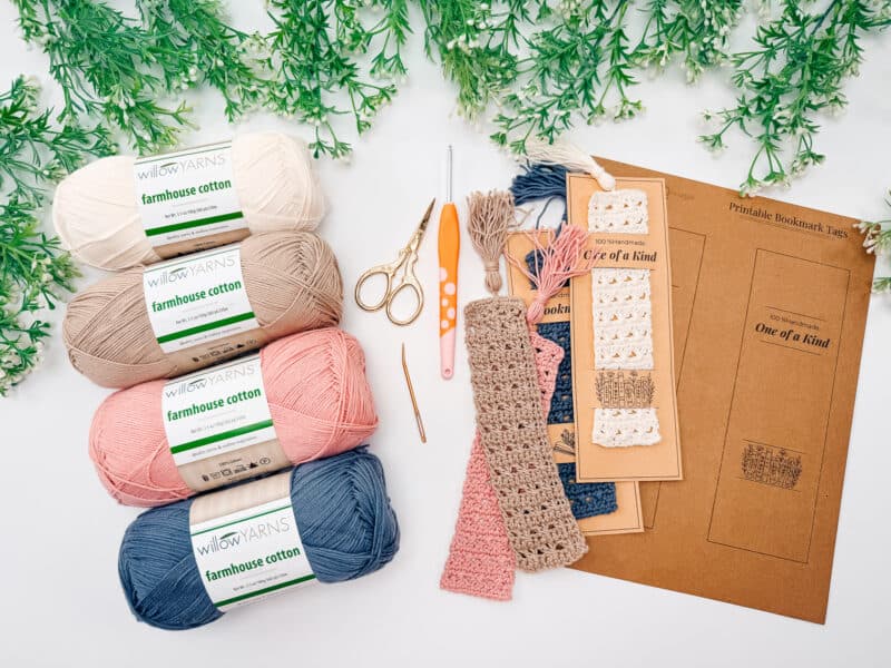 Four skeins of cotton yarn, crochet tools, crocheted bookmarks, and printable tags are arranged on a white surface with green foliage at the top.