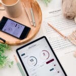 A tablet and smartphone display a stitch tracking app on a table with crochet hooks, yarn, a cup of coffee, and a printed stitch chart surrounded by green plants.