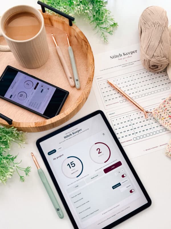 A tablet and smartphone display a stitch tracking app on a table with crochet hooks, yarn, a cup of coffee, and a printed stitch chart surrounded by green plants.