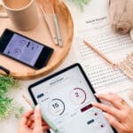 A person uses a tablet with a "Stitch Keeper" app, surrounded by crochet supplies, a printed pattern, a phone, a cup of coffee, and yarn on a table.