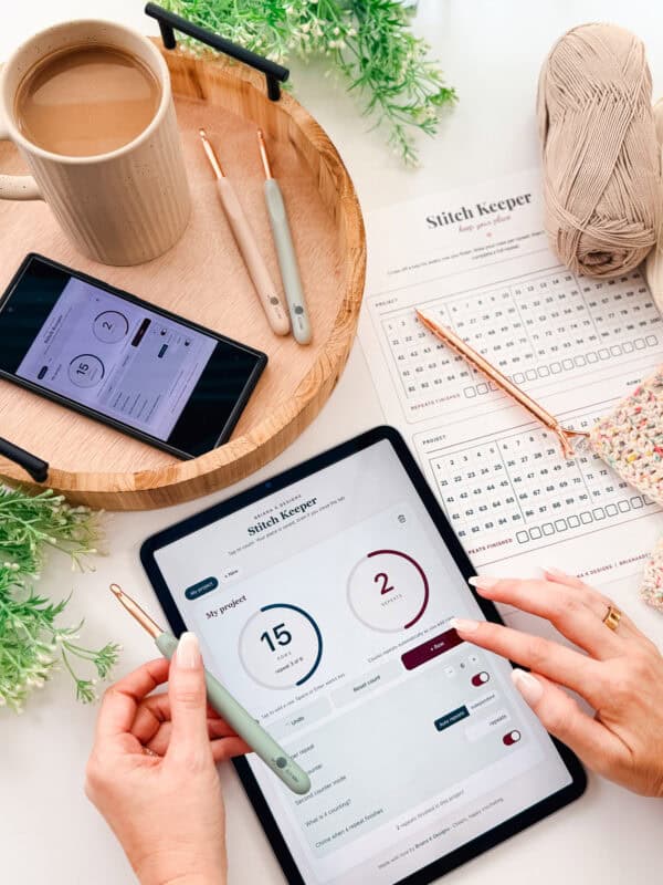 A person uses a tablet with a "Stitch Keeper" app, surrounded by crochet supplies, a printed pattern, a phone, a cup of coffee, and yarn on a table.
