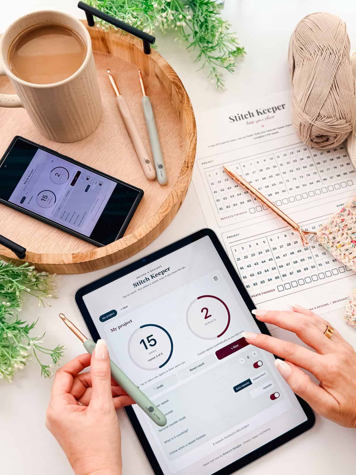 A person uses a tablet with a "Stitch Keeper" app, surrounded by crochet supplies, a printed pattern, a phone, a cup of coffee, and yarn on a table.