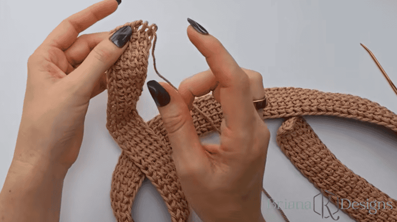 Two hands hold and crochet a strip of pink yarn using a crochet hook, with several finished crochet pieces visible on a white surface.