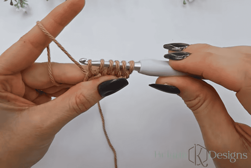 Close-up of hands holding a crochet hook and working with light brown yarn, forming multiple loops on the hook.