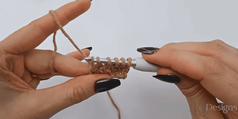 Close-up of two hands holding a crochet hook and working with light brown yarn, creating several loops on the hook against a white background.