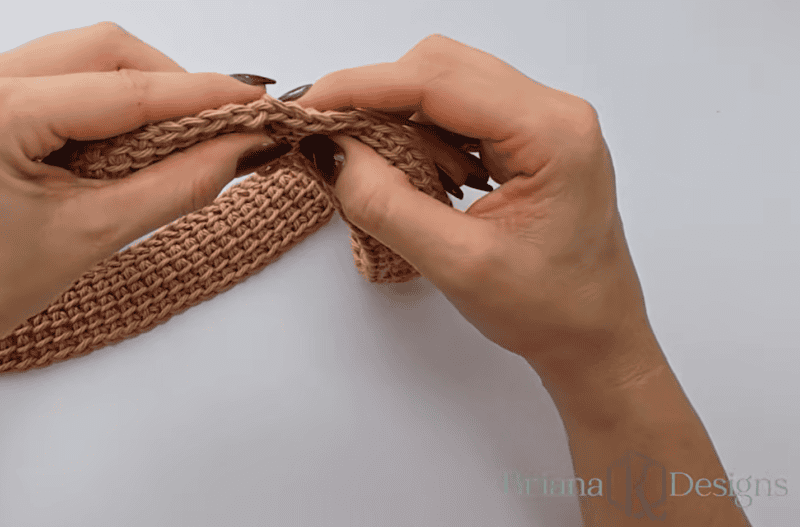 Two hands with black nail polish hold and fold a pink crocheted strip against a white background.