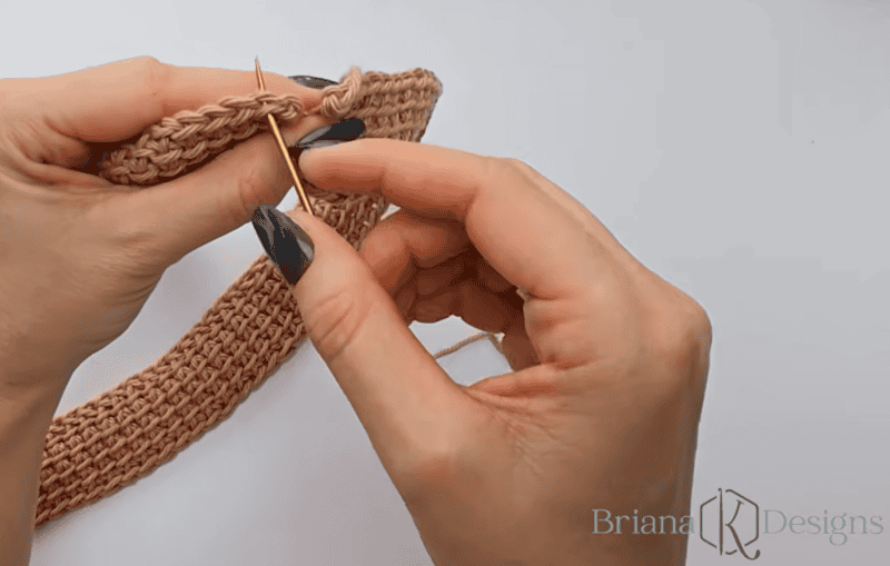 Two hands with dark nail polish use a yarn needle to sew together the edges of a light brown crocheted fabric piece on a white surface.