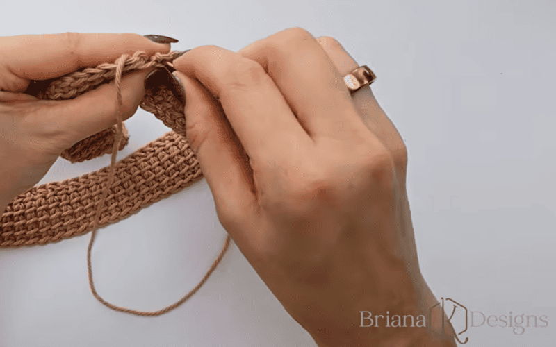 A person crocheting with light brown yarn, holding a crochet hook and working on a textured piece against a white background.
