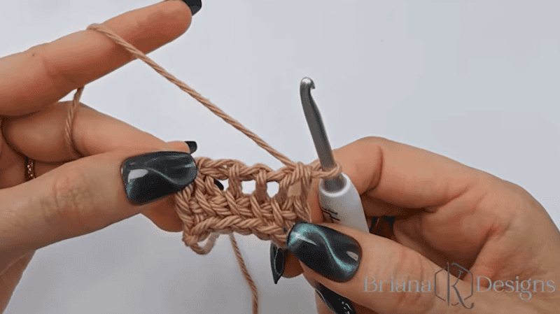 Two hands hold a crochet swatch in progress with beige yarn and a metal crochet hook against a white background.