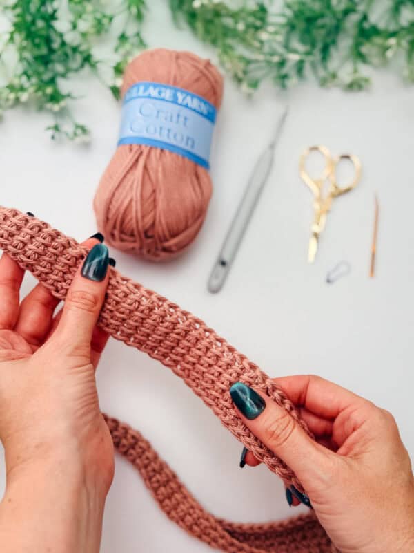 Hands holding a pink crochet piece with a ball of yarn, crochet hook, scissors, needle, and stitch marker on a white surface in the background.
