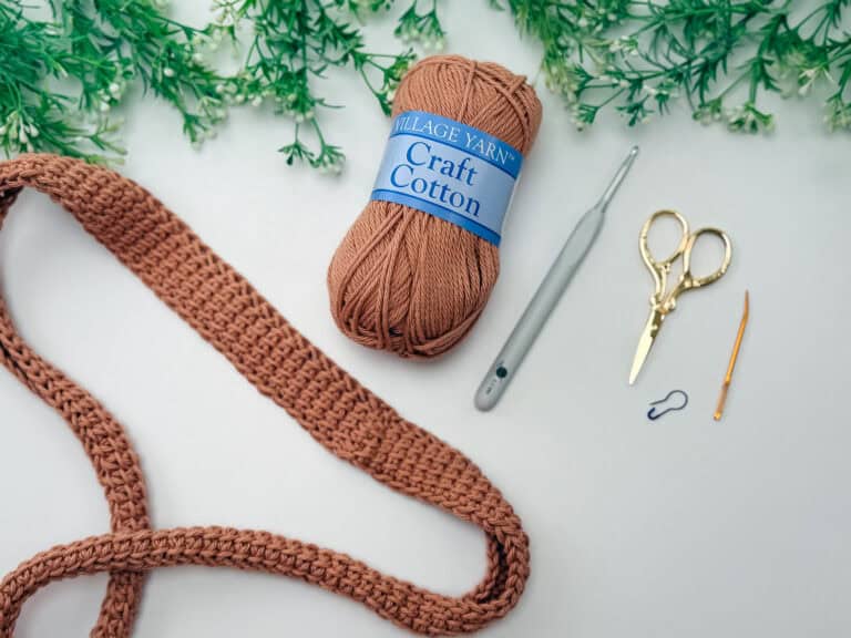 A ball of brown craft cotton yarn, a crochet hook, scissors, a tapestry needle, a stitch marker, and a crocheted project on a white surface with green foliage at the top.