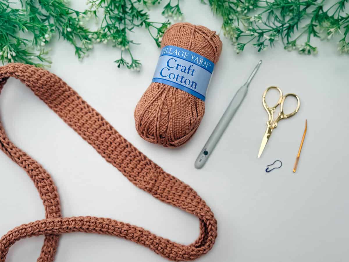 A ball of brown craft cotton yarn, a crochet hook, scissors, a tapestry needle, a stitch marker, and a crocheted project on a white surface with green foliage at the top.