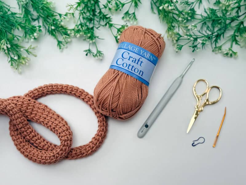 Brown cotton yarn, a crochet hook, scissors, a yarn needle, a stitch marker, and a crocheted piece are arranged on a light surface with green foliage at the top.