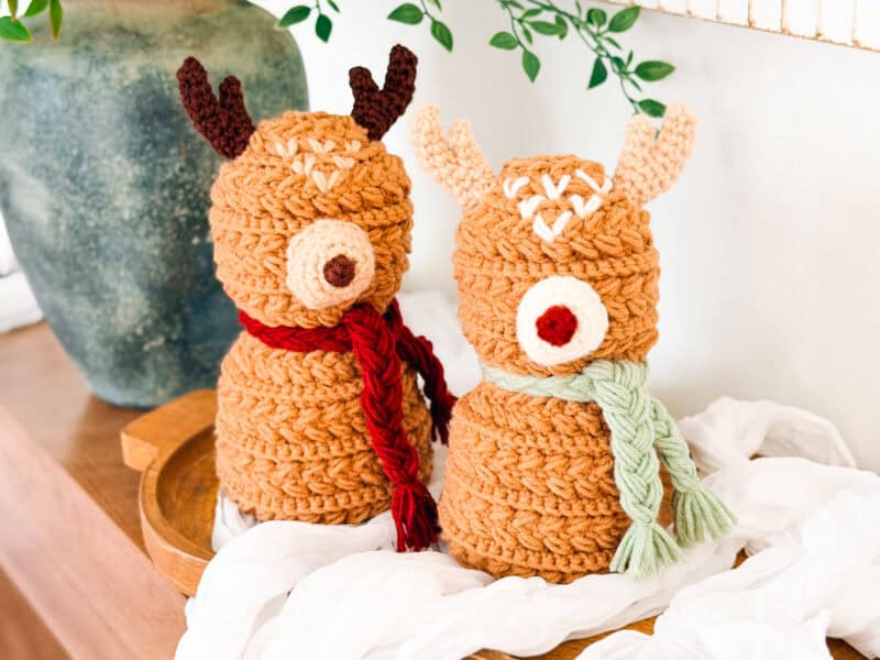 Two crocheted reindeer figures with scarves stand side by side on a wooden tray, with a plant and white fabric in the background.