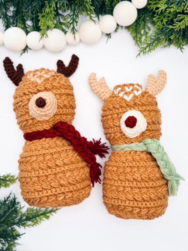 Two crocheted reindeer dolls with scarves, one red and one green, are displayed on a white background with greenery and white decorative beads.