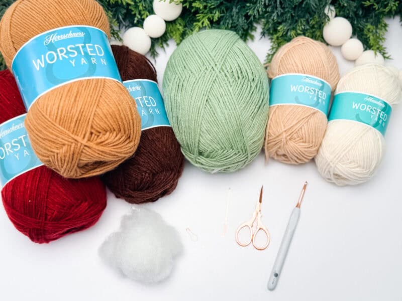 Several skeins of yarn in red, brown, green, beige, and white, with crochet tools, fiber fill, and greenery arranged on a white surface.