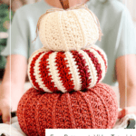 A person holds a wooden tray with three crocheted pumpkins stacked on top of each other, in cream, striped red and white, and orange colors.