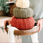 A person holds a wooden tray with three crocheted pumpkins in cream, green, and orange, stacked by size. Text at the bottom reads: “Petal Loop Pumpkin Trio - 3 Sizes.”.