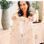 Woman holding a large, off-white, crochet mesh bag with a knot at the top. Text above reads “Knotted Mesh Bag Crochet Pattern + Video Tutorial.”.