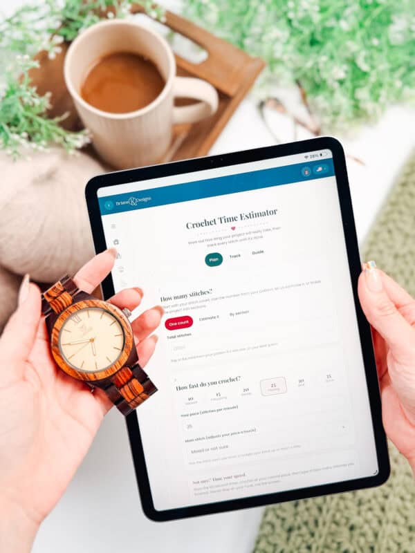 Person holding a wooden wristwatch and an iPad displaying a crochet time estimator page with a yarn stash tracker; a cup of coffee and green plants are in the background.
