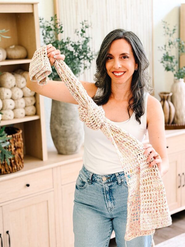 A woman in a white tank top and jeans holds up a crocheted scarf in a room with shelves, plants, and decorative vases.