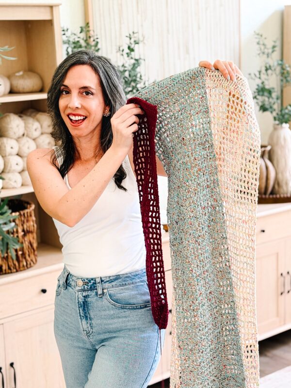 Woman in a white tank top and jeans holds up a crocheted blanket with a maroon border, standing in a room with shelves of yarn and decorative plants.
