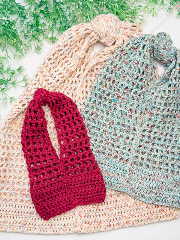 Three crocheted market bags in beige, blue, and red are displayed on a white surface, with green decorative foliage at the top.
