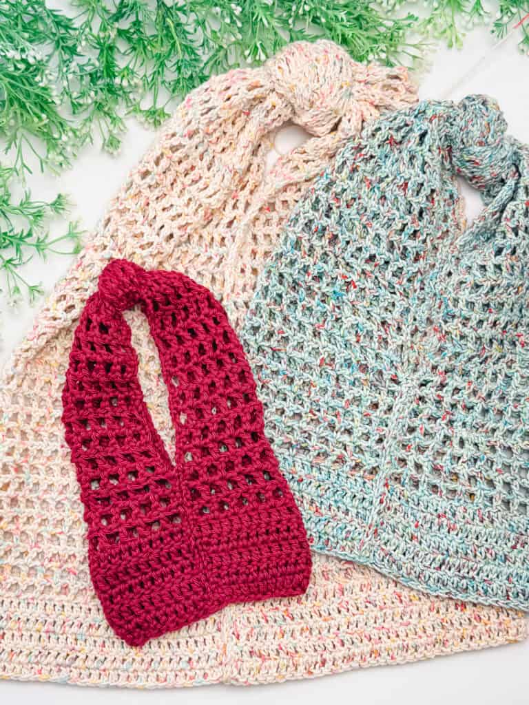 Three crocheted market bags in beige, blue, and red are displayed on a white surface, with green decorative foliage at the top.