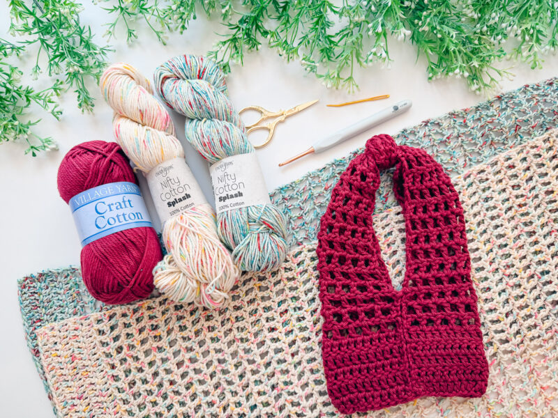 Three yarn skeins, crochet hooks, scissors, and a finished maroon crochet headband are arranged on a multicolored crocheted mat with green foliage in the background.