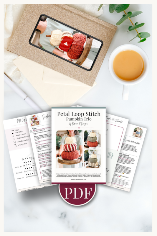 Flat lay of a printed crochet pumpkin pattern, instruction sheets, a PDF icon, a cup of coffee, an envelope, and a tablet displaying a photo of crocheted pumpkins.