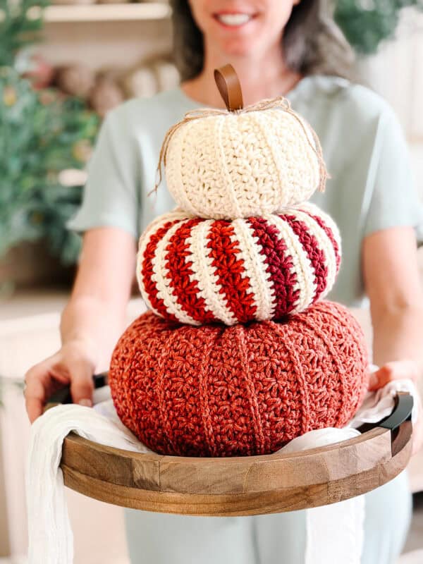A woman holds a wooden tray with three stacked crocheted pumpkins in orange, striped red and white, and cream colors.